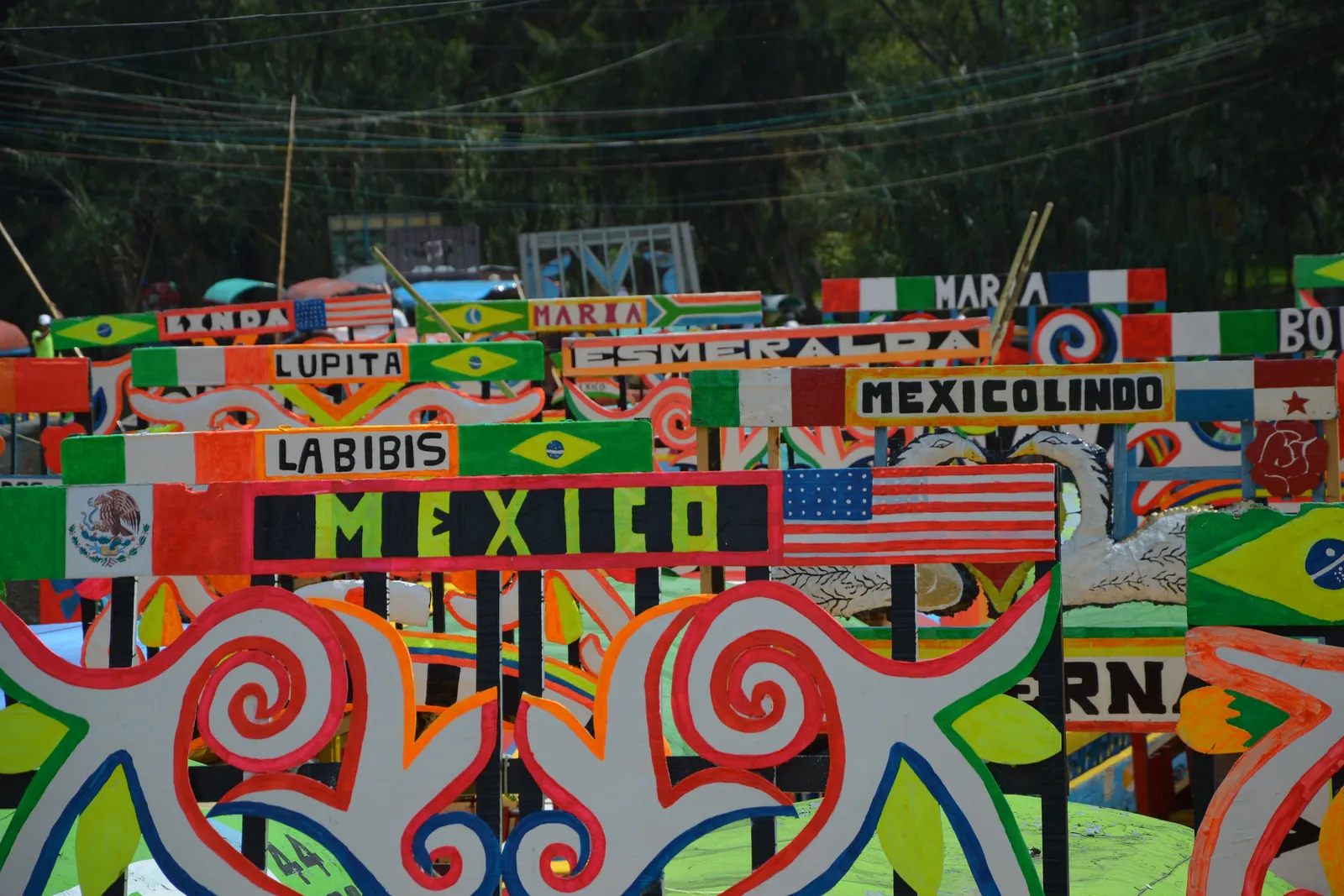 Close view of trajinera name arches at a Xochimilco landing, painted with flowers, flags and women's names