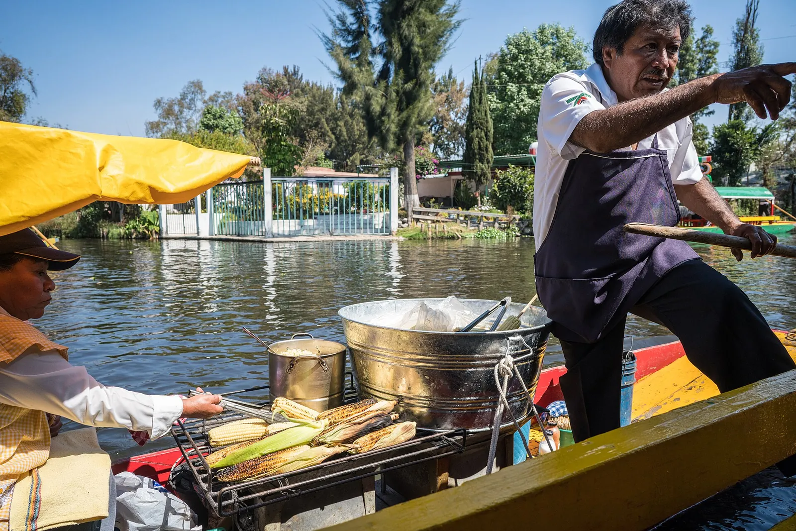 A boatman standing at the stern of a small boat in a Xochimilco canal, grilling corn on a charcoal grill