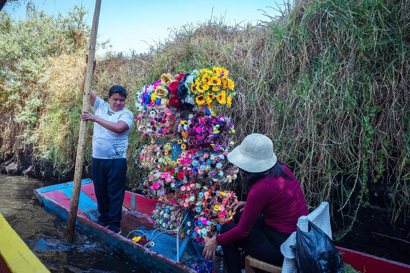 A vendor boat in a Xochimilco canal loaded with bunches of artificial flowers and flower crowns, with a boatman standing at the stern