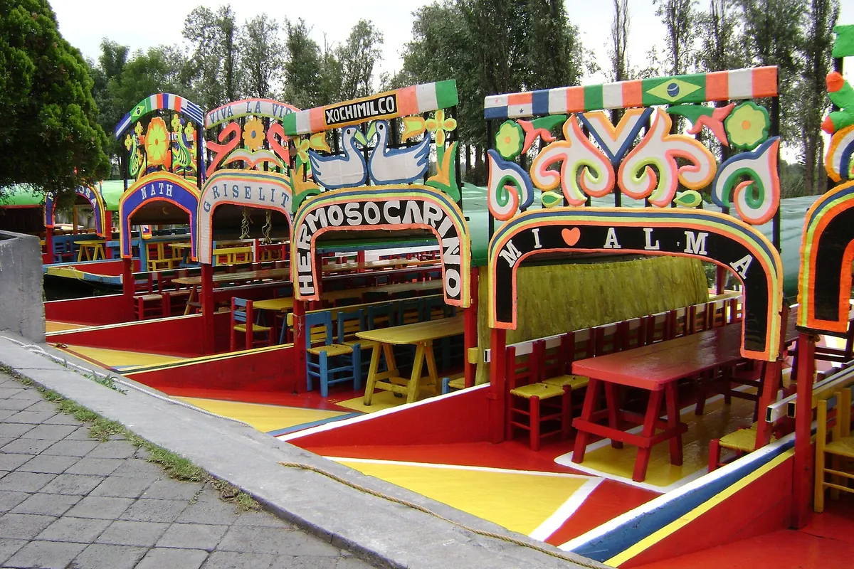 Painted trajineras with arched name boards and wooden tables tied up at the Cuemanco landing in Xochimilco