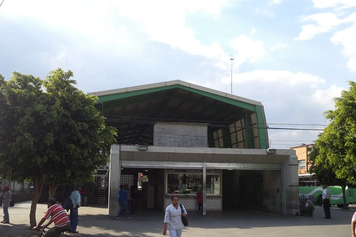 The entrance building of a light rail station in Xochimilco, with a green roof, a ticket office and people walking past under trees