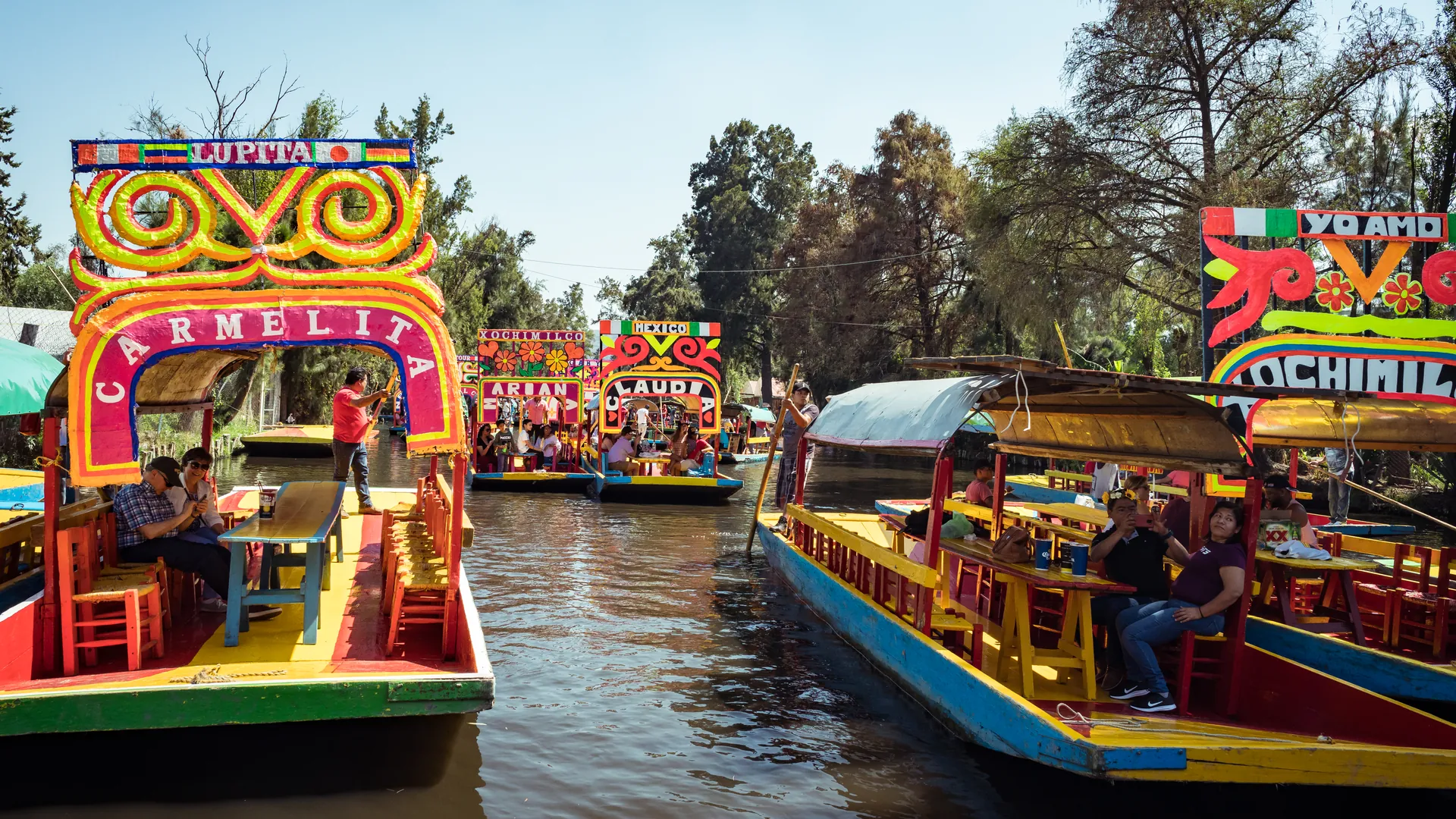 Brightly painted trajineras moored side by side at a Xochimilco landing, their arched name boards in red, yellow and green, with a dark canal to the left and trees beyond