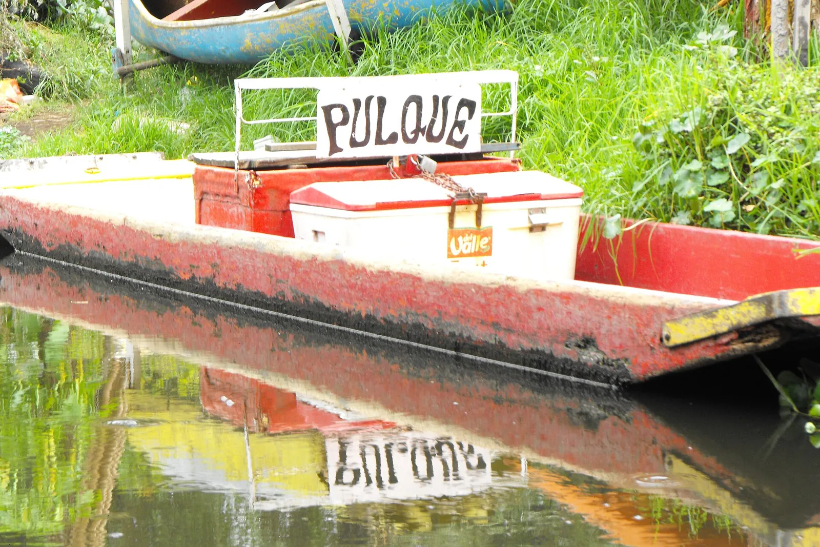 A red boat on a Xochimilco canal carrying a cooler and a hand-painted sign reading pulque